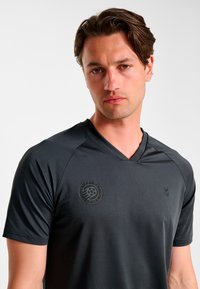 Czarna sportowa koszulka z dekoltem w serek, krótkimi rękawami, teksturowaną tkaniną, z subtelnym logo na piersi i mniejszym emblematem na rękawie.