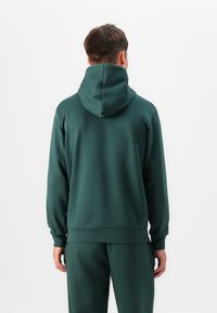 Felpa verde realizzata in tessuto morbido, con un ampio cappuccio, polsini e vita a costine. Design semplice senza modelli o elementi hardware.