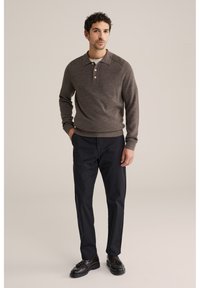 Hombre con una camisa de polo de manga larga de punto marrón, pantalones negros y mocasines negros con borlas, de pie con una mano en el bolsillo.