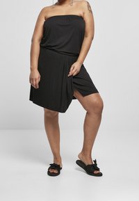 Nainen mustassa hihattomassa mekossa seisoo jalka hieman koukistettuna, jalassaan mustat liukuvaellussandaalit, joissa on nauhat, harmaan taustan edessä.