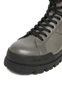 Pelēks ādas zābaks ar melnām šņorēm, ar teksturētu gumijas zolīti un izteiktu logotipu. Apaļa pirksta daļa un pastiprināta pirksta daļa ilgmūžībai.