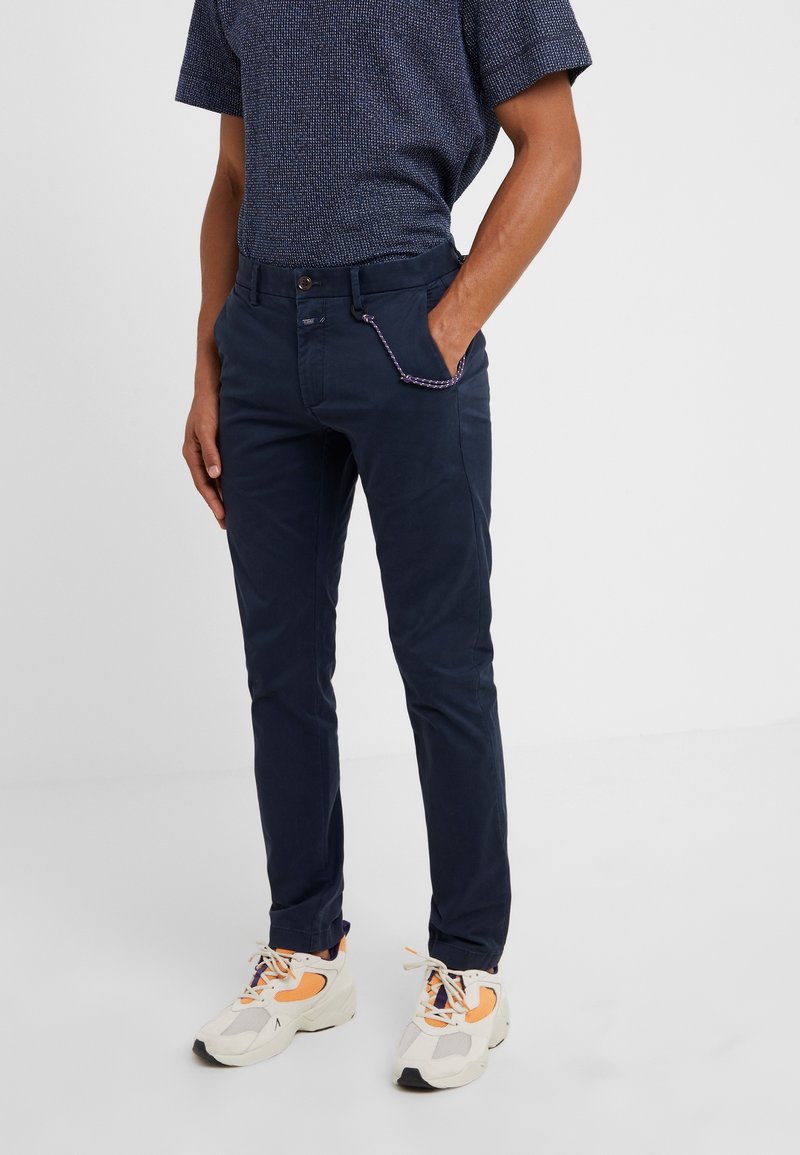 Homme portant un pantalon slim marine avec une chaîne à détail corde, une chemise texturée à manches courtes marine, et des baskets blanches avec des accents orange.