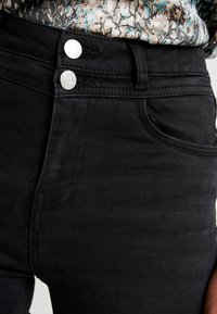 Los jeans de mezclilla negros tienen una cintura alta, cierre de doble botón y bolsillos frontales. La textura es suave con detalles de costura sutiles.