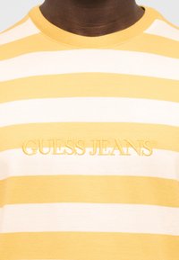 Κίτρινη και λευκή οριζόντια ριγέ μπλούζα με κεντημένο το "GUESS JEANS" στο στήθος, φορεμένη από ένα άτομο με σκουρόχρωμο δέρμα.