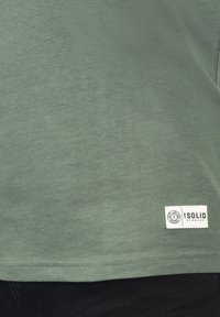 Grøn bomulds t-shirt med en glat tekstur, der har en lille hvid logoetiket nederst til højre. Enkel design med rund hals.