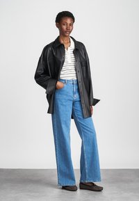 Μαύρο δερμάτινο overshirt, ριγέ κοντομάνικο μπλουζάκι, φαρδιά μπλε τζιν παντελόνια και καφέ slip-on παπούτσια. Απλός σχεδιασμός με δομημένη εφαρμογή.