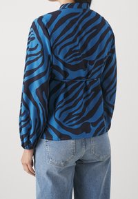 Blouse à motifs bleus et noirs avec manches longues, dos plissé et col arrondi, confectionnée dans un tissu lisse et léger.