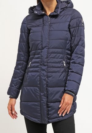 Femme portant une longue doudoune matelassée bleu marine avec capuche, fermeture éclair frontale, et poches latérales zippées, debout devant un fond uni.