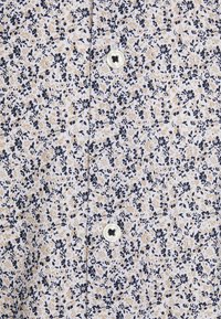 Lichtgekleurde bloesemshirt met een marineblauw en gouden patroon, voorzien van een ronde witte knop en een gestructureerde stof.