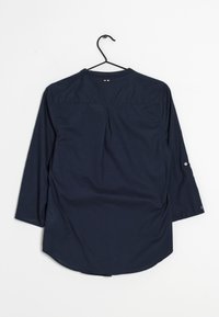 Marinblå långärmad skjorta med knappmanschetter och en liten röd, vit och svart broderad detalj på övre ryggen hängande på en svart galge.