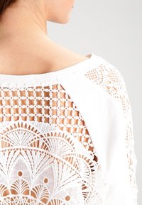 Spalla e parte superiore della schiena di una donna visibili, che indossa un top bianco con dettagli intricati in pizzo all'uncinetto e motivi geometrici.