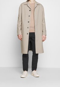 Manteau à carreaux beige avec un col pointu, des boutons à l'avant et deux poches avant, associé à un pull beige, un pantalon à rayures et des baskets blanches.