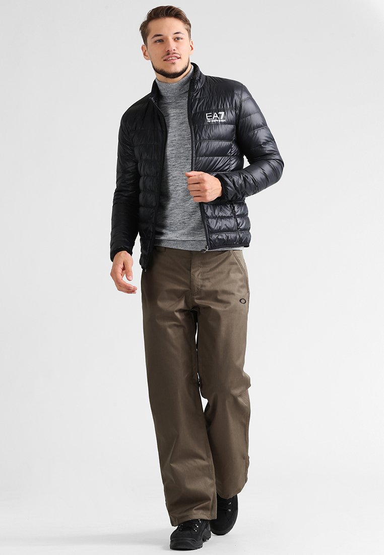Jachetă puf cu design pătrățit orizontal, de culoare neagră, asociată cu un pulover gri cu guler înalt, pantaloni maro și încălțări negre. Un aspect simplu și funcțional.