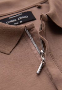 Camisa polo marrón con cremallera y tejido texturizado, presenta una cremallera metálica y una etiqueta etiquetada como "OMBRE EXIMO". Primer plano del detalle del collar y la cremallera.