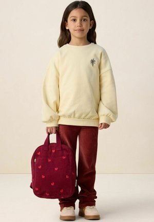 Fille aux longs cheveux bruns portant un sweat-shirt jaune pâle avec un petit palmier, un pantalon en velours côtelé couleur rouille, tenant un sac à dos rouge à motifs de cœurs.
