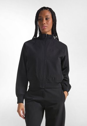 Woman with long braided hair wearing a black zip-up jacket and matching black pants, standing with one hand in pocket against a white background.
