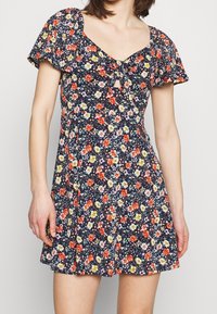 Vestido floral azul marino con mangas cortas acampanadas, que presenta pequeñas flores rojas, amarillas y moradas, un detalle de nudo en el frente y una cintura entallada.