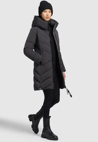 Zwarte pufferjas met een capuchon, voorzien van een chevron-gestikt ontwerp en ritssluiting. Gecombineerd met zwarte leggings en enkellaarsjes.
