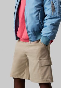 Giacca bomber azzurro chiaro, felpa rossa sotto, e pantaloni cargo beige con tasche. Il tessuto appare liscio e di design casual.