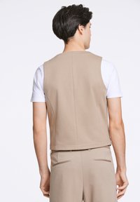 Beige ärmlös väst i slät tyg. Har en ren design med raka kanter och inga utsmyckningar, bärs över en vit skjorta.