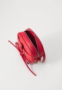 Sac en bandoulière en cuir rouge avec une texture lisse, une forme arrondie, une fermeture zippée et une sangle réglable. Avec des accents métalliques et un détail en nœud.