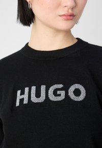 Femme aux cheveux courts et foncés, portant des boucles d'oreilles argentées en forme d'anneau et un pull noir avec le mot « HUGO » tricoté en blanc sur la poitrine.
