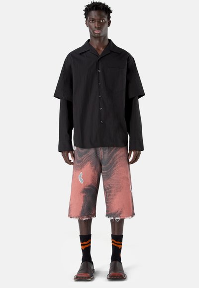 Hombre con camisa negra de manga larga holgada combinada con una camiseta de manga corta, pantalones cortos hasta la rodilla con patrón rosa y negro, calcetines negros y zapatos negros.