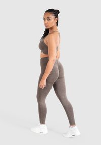 Conjunto de sujetador deportivo y leggings marrones, hecho de una tela suave y elástica con una textura lisa. La modelo posa de lado, llevando zapatillas blancas.