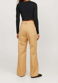 Beige weitgeschnittene Hose mit hoher Taille und Gesäßtaschen. Mit einem Logo über der rechten Tasche. Kombiniert mit einem schwarzen, cropped Langarmoberteil.