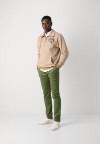 Beżowa bluza zapinana na zamek z logo, zielone spodnie i białe sneakersy. Gładki materiał, luźny krój, z warstwowym wyglądem spowodowanym koszulą z kołnierzykiem pod spodem.