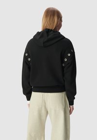 Felpa nera con cappuccio regolabile, dettagli con occhielli sulle maniche, polsini e orlo a coste. Abbinata a pantaloni chiari a gamba ampia.