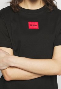 T-shirt noir à manches courtes avec un patch rectangulaire rouge affichant "HUGO" en texte noir au centre. Matière en coton, design simple.