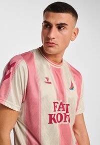 Koszulka z krótkim rękawem w jasnobeżowym kolorze z różowymi pionowymi paskami. Zawiera napis „FAT KONT” w wyrazistej czerwieni, logo Hummel oraz wielokolorowy akcent na kołnierzu.