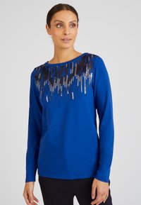 Maglia a maniche lunghe blu con design di paillettes sul collo, accompagnato da dettagli metallici neri e argento. Tessuto morbido e vestibilità rilassata.