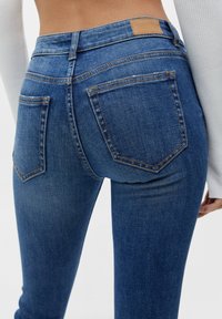 Jeans ajustados en denim azul oscuro con una textura suave, que cuentan con un parche de cuero marrón en la cintura y dos bolsillos traseros con detalles de costura.