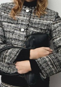 Czarna i biała marynarka w jodełkę z srebrnymi guzikami i czarnymi mankietami. Trzyma małą czarną skórzaną torbę o fakturowanej powierzchni i zamku błyskawicznym.