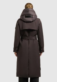 Donna con bun biondo che indossa un lungo cappotto invernale marrone scuro con cintura e colletto alto imbottito, vista di spalle su uno sfondo chiaro e uniforme.