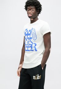 Biele bavlnené tričko s modrým grafickým textom a motívmi, spojené s čiernymi teplákmi s logom "nike" v zlatej farbe, voľný strih.