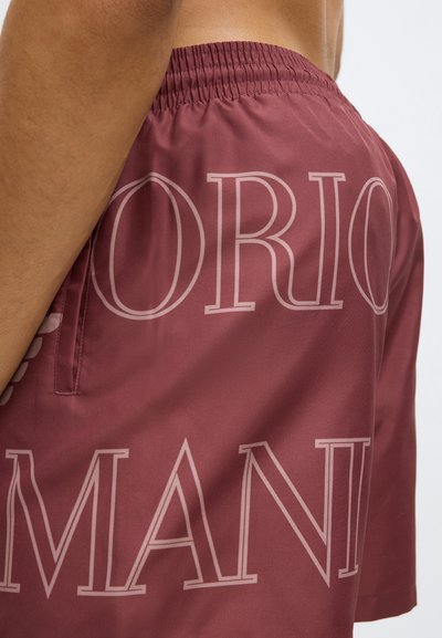 Bordó úszónadrág sima anyagból; elasztikus derékrésszel és világosabb árnyalatú nagy szövegnyomattal. Oldalzsebbel ellátva.