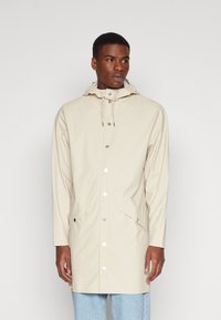 Lys beige regnfrakk med hette, trykknapplukking og sidelommer. Glatt tekstur, lange ermer og rett snittdesign.