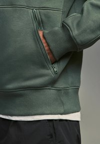 Sudadera verde con dobladillo y puños acanalados, que presenta un bolsillo lateral con cremallera. El material parece suave y texturizado.