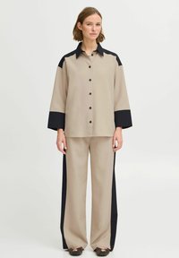 Cămașă bej cu nasturi și accente negre, asociată cu pantaloni largi bej, cu panouri laterale negre. Textură netedă, ajustare pe măsură.