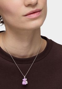 Fioletowy wisiorek w kształcie niedźwiedzia na srebrnym łańcuszku, na tle ciemnej koszuli. Niedźwiedź ma błyszczące wykończenie i mały biały akcent.