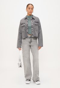Chaqueta de mezclilla con cierre de botones, dos bolsillos frontales y un diseño corto en gris, combinada con una blusa verde y jeans claros de tiro alto.