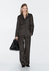 Donna che indossa un abito scuro a righe con blazer abbottonato e pantaloni a gamba larga, mentre tiene una grande borsa nera con texture, in piedi su uno sfondo neutro.