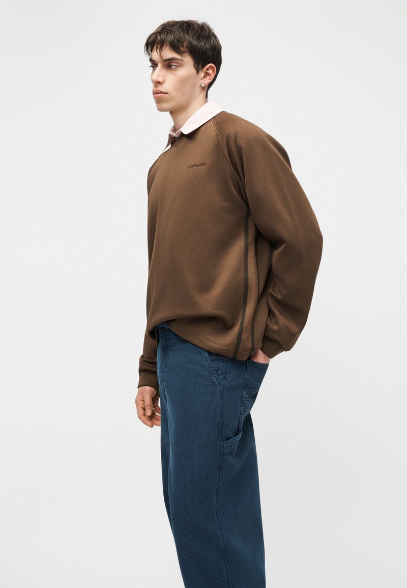 Jeune homme portant une chemise marron à manches longues par-dessus une chemise blanche à col, et un pantalon bleu, debout avec une main dans la poche, regardant sur le côté.