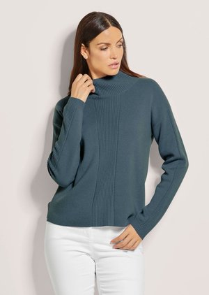 Vrouw draagt een teal geribbelde coltrui en witte broek, houdt de kraag met één hand vast en kijkt opzij.