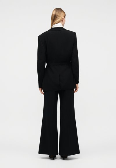 Γυναίκα με μακριά ξανθά μαλλιά, γυρισμένη με την πλάτη, φοράει μαύρο σακάκι και μαύρο παντελόνι με φαρδιά μπατζάκια, σε απλό φόντο.