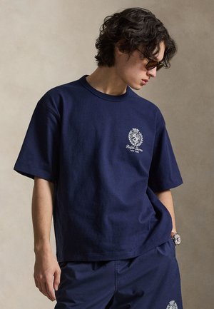 Νέος άνδρας που φορά μπλε Ralph Lauren T-shirt και αντίστοιχα σορτς, κοιτάζοντας προς τα κάτω, με γυαλιά ηλίου και ρολόι χειρός.
