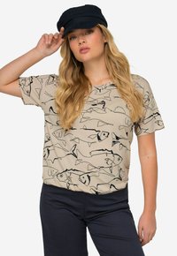 Camiseta beige con un patrón de peces negros, mangas cortas y cuello redondo, combinada con pantalones azul marino oscuros y una gorra negra.
